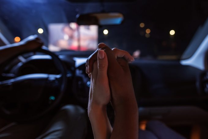 Close up of diverse young couple holding their hands together while having a romantic evening. Young people watching a movie at drive in cinema from the front seats of the car