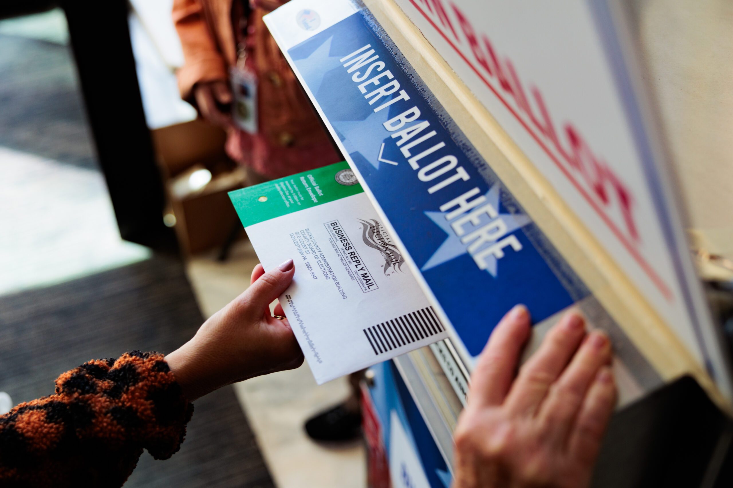 Ballot envelope inserted into voting box, marked "INSERT BALLOT HERE," amidst an indoor voting setting.