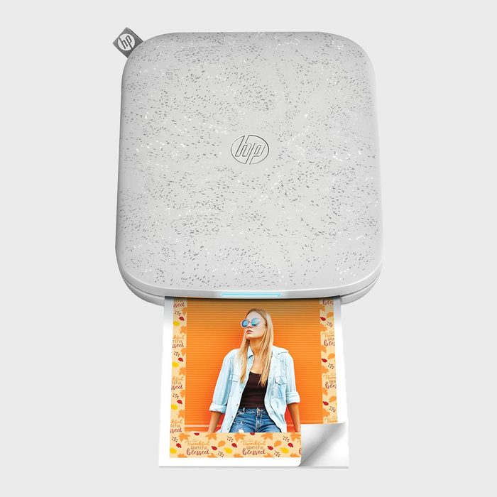 A small white printer ejects a colorful photo of a woman against an orange background, set in a floral border, on a plain surface.