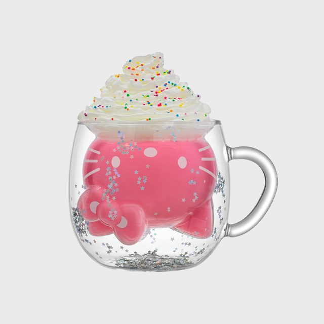 A transparent mug contains pink liquid, topped with whipped cream and colorful sprinkles, surrounded by silver star confetti on a plain background.