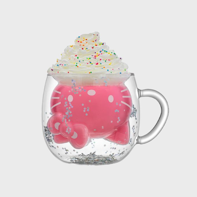 A transparent mug contains pink liquid, topped with whipped cream and colorful sprinkles, surrounded by silver star confetti on a plain background.