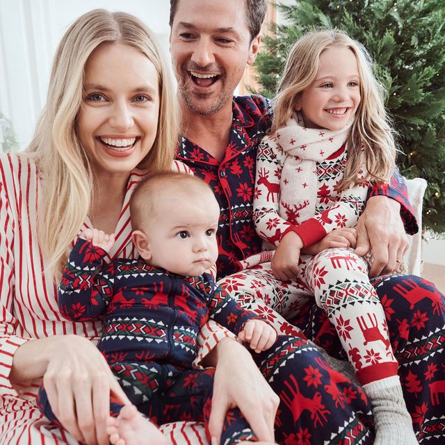 Family sits together, wearing matching festive pajamas, smiling in a cozy room with a Christmas tree in the background.
