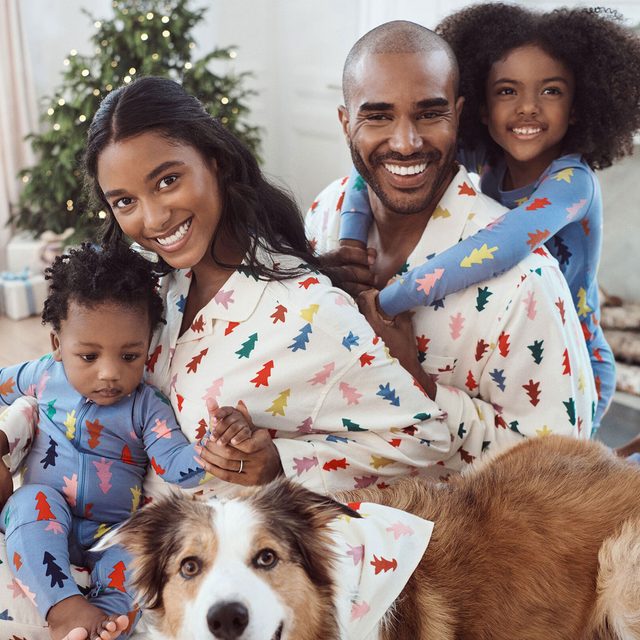 Family smiles and sits together in matching tree-patterned pajamas, indoors near a decorated Christmas tree with a dog in a similar outfit.
