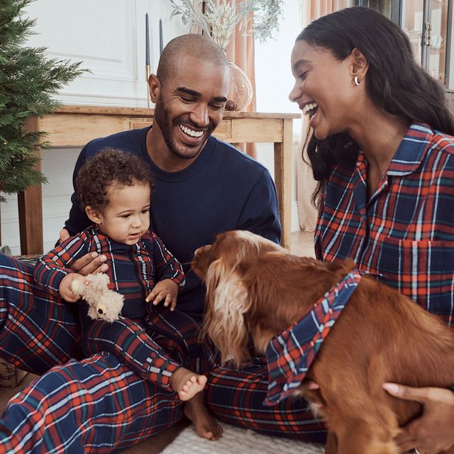 A family, wearing matching plaid pajamas, sits with a child and dog, sharing smiles and laughter in a cozy indoor setting.