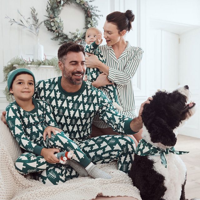 Family wearing matching tree-patterned pajamas smiles while sitting indoors, petting a large black-and-white dog, with holiday wreaths and dÃ©cor around.