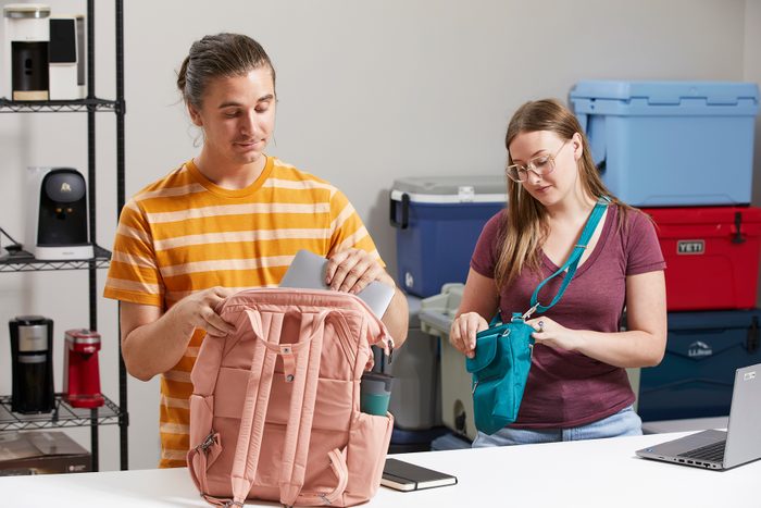 A person places items into a pink backpack, while another organizes a teal bag, in a room with stacked coolers and shelves.