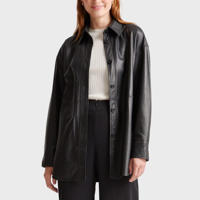 A person wears a black leather jacket over a white sweater, standing against a plain gray background.