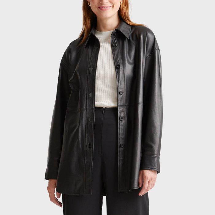 A person wears a black leather jacket over a white sweater, standing against a plain gray background.