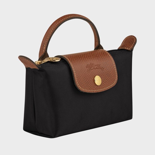 A black handbag with a brown leather flap and handle, features a gold button, set against a plain gray background.