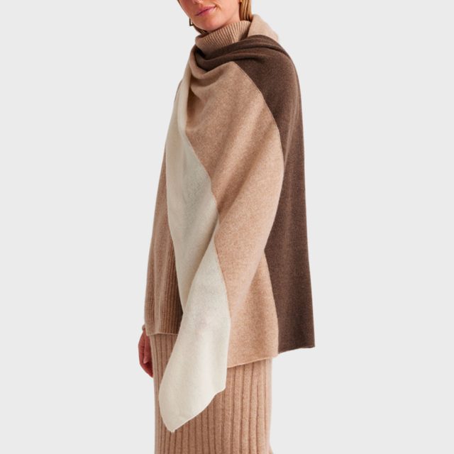 Woman wears a draped, color-blocked poncho in brown, beige, and white, standing against a plain, light background.