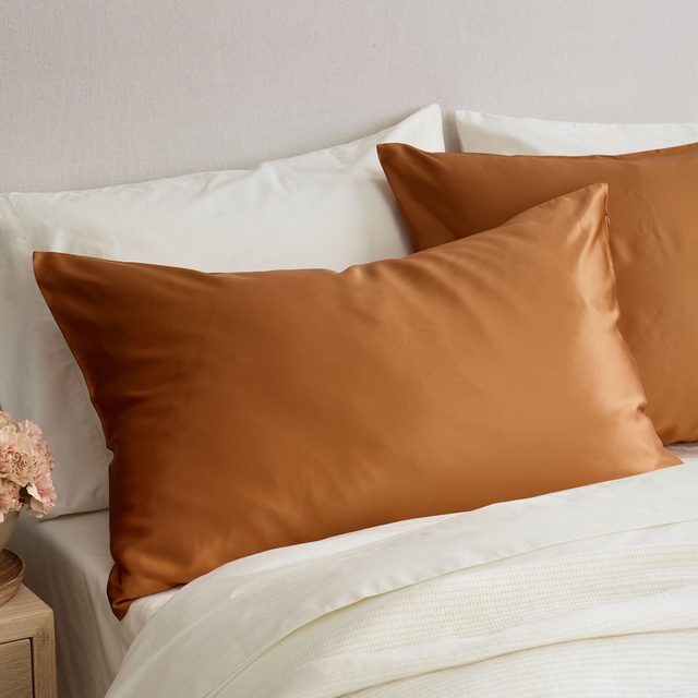 Copper-colored silk pillow rests on white bed linens, set against a light-colored backdrop with pink flowers on a wooden nightstand.