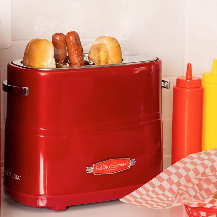 Retro toaster cooks hot dogs and buns; red and chrome design. Nearby, red and yellow condiment bottles and patterned napkins on countertop.