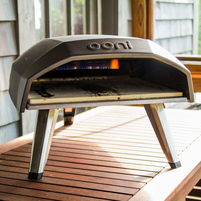 Ooni pizza oven sits on a wooden table, with flames visible inside, surrounded by a wooden-paneled outdoor patio.