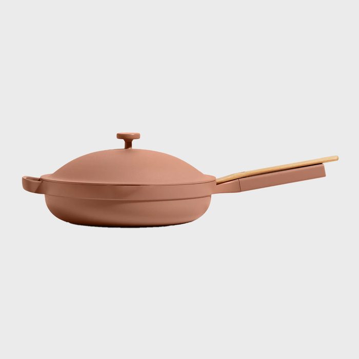 A terracotta-colored pan with a lid rests on a neutral background, featuring a wooden handle extended horizontally.