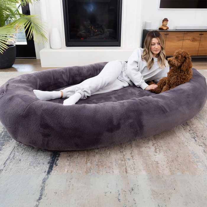 Woman lounges on a large, plush dog bed, interacting with a brown dog, in a cozy living room with fireplace and plants.