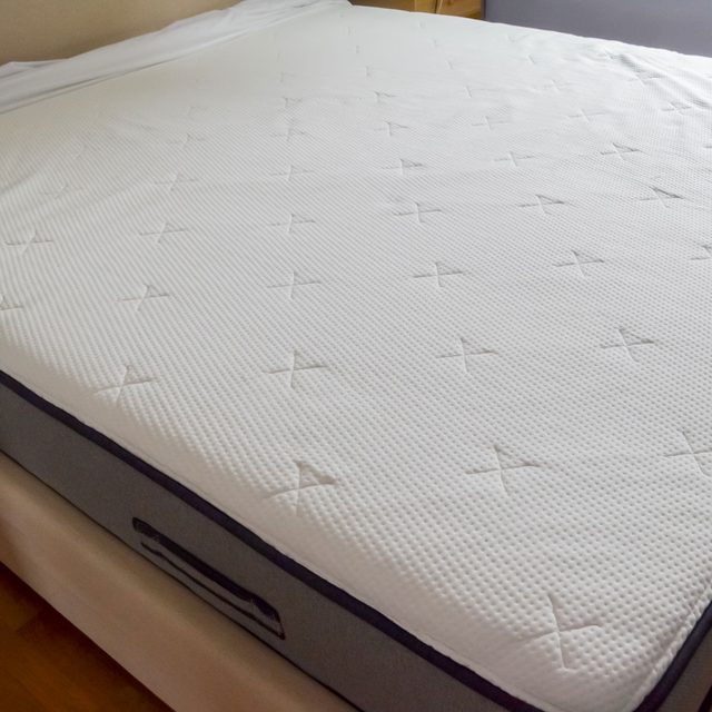 Mattress rests on a bed frame, featuring a textured white surface with cross patterns, in a bedroom setting with wooden floors.
