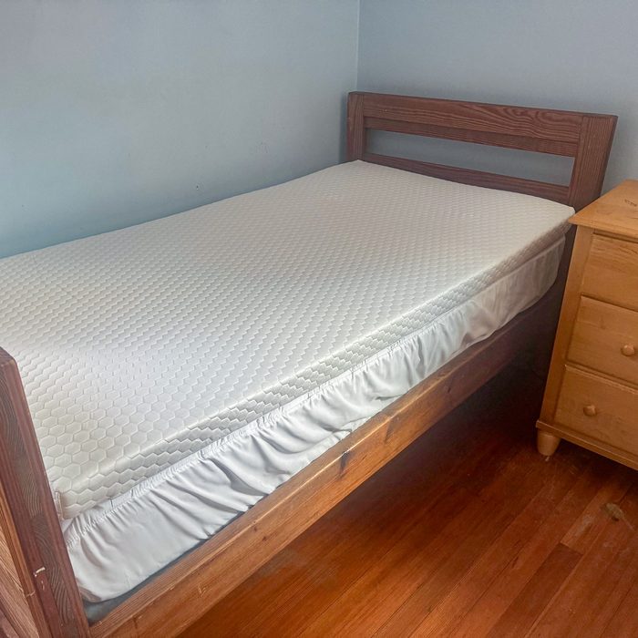 Bed sits unoccupied, covered in white quilted mattress protector; wooden floors and dresser fill the adjacent bedroom space.