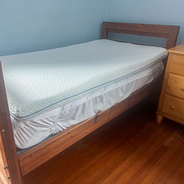 A neatly-made, twin-sized bed sits against a blue wall, beside a wooden nightstand on a polished wooden floor.