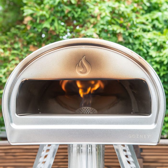 Pizza oven emits flames outdoors, surrounded by lush green foliage.