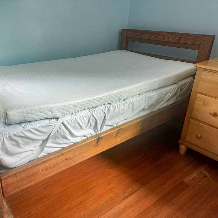 Bed with white mattress resting on wooden frame beside a light wood dresser in a room with light blue walls.