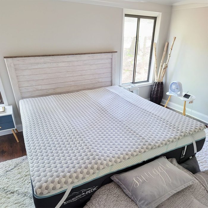 Mattress with textured topper lies on a bed in a well-lit bedroom. Nearby, a fan and small table sit beside a window.