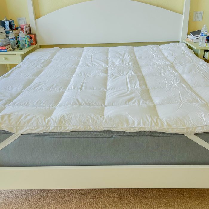 A bed with a white mattress topper, flanked by matching nightstands cluttered with books and water bottles in a carpeted bedroom.