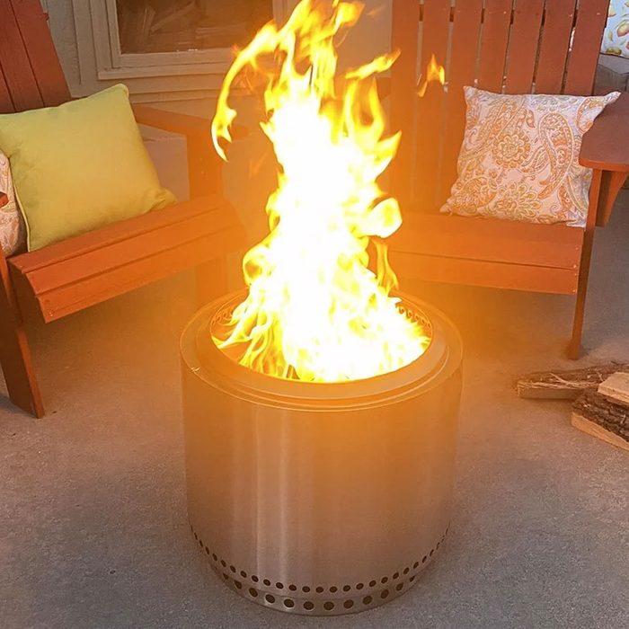 A metal fire pit emits tall flames, surrounded by wooden chairs with colorful cushions, set on a concrete patio.