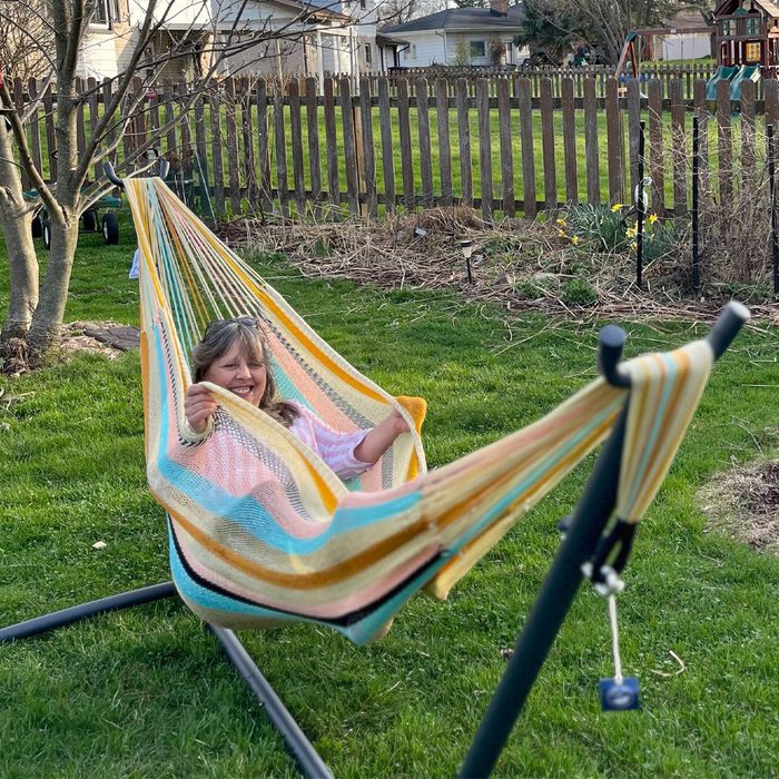 Woman reclining comfortably in a striped hammock, smiling in a grassy backyard, bordered by a wooden fence and leafless trees.