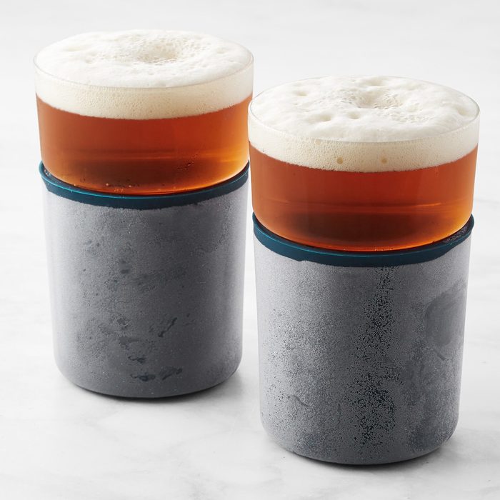 Two glasses hold amber liquid with foam, resting inside matte silver holders on a smooth, white surface.