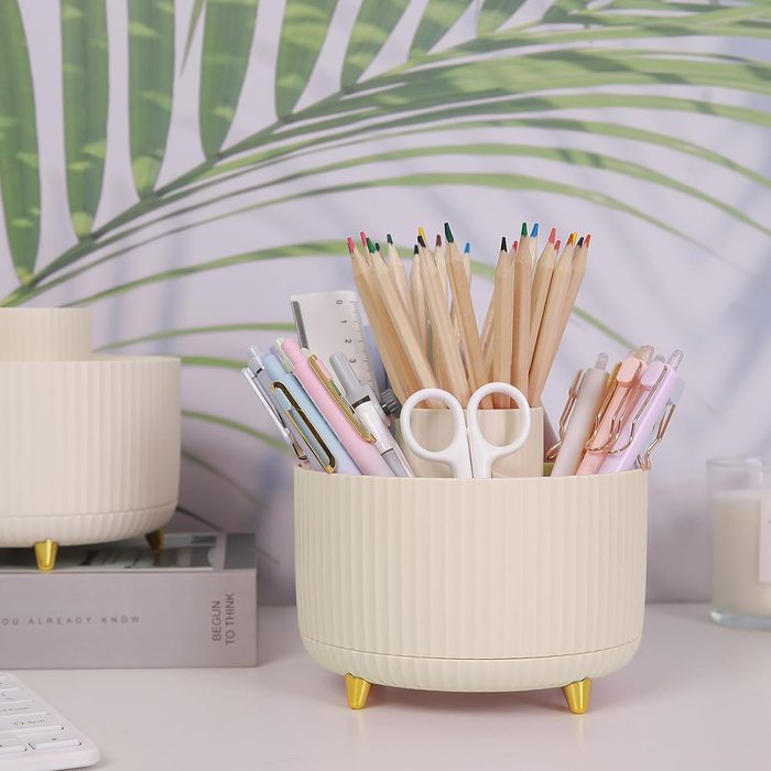 A cream-colored organizer holds pencils, pens, and scissors, situated on a desk with leafy wallpaper in the background. Nearby, a book reads 