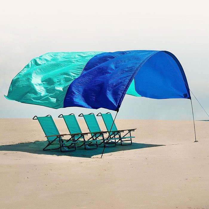 Four beach chairs sit under a large, colorful canopy, providing shade on a sandy beach with a clear sky above.