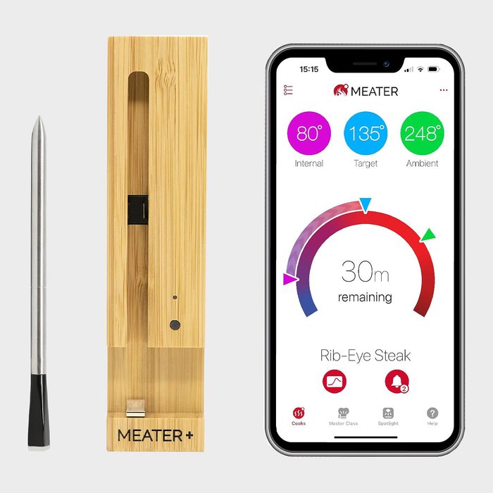 Thermometer displays temperature data on a smartphone app, showing cooking progress for Rib-Eye Steak with a wooden charging base nearby, in a kitchen setting.