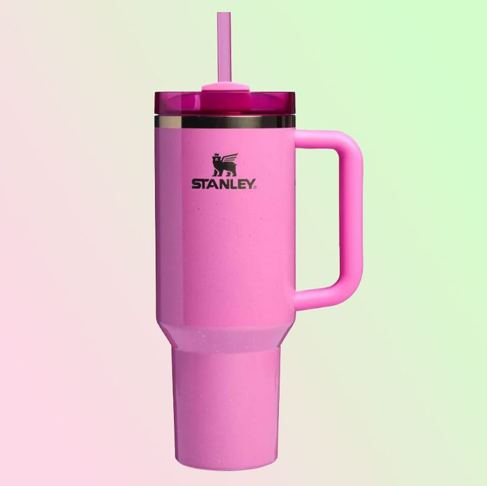 A pink insulated tumbler with a handle and straw stands against a pastel gradient background. Logo reads â€œStanleyâ€.