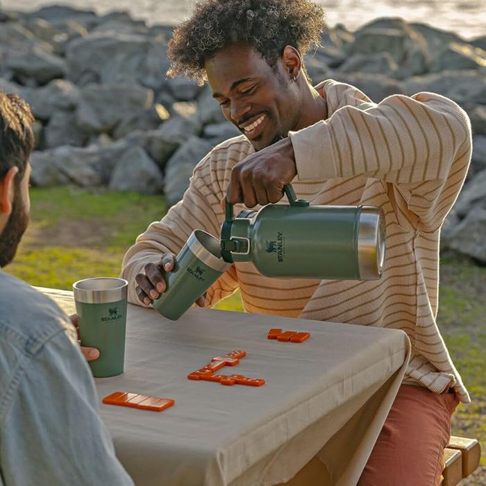 Man pours from green thermos into cup, sitting at table with orange tiles, outdoors near rocky shoreline.