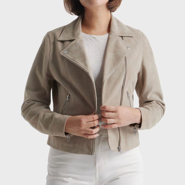 Person adjusts a beige suede jacket while standing, against a plain white background.