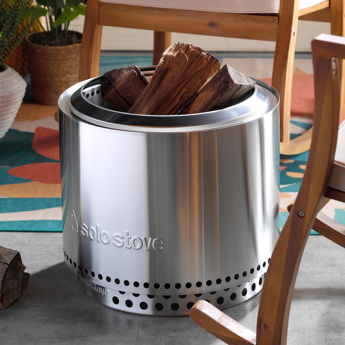 Metal stove holds logs, positioned on a colorful rug, surrounded by wooden chairs and potted plants. Text reads 