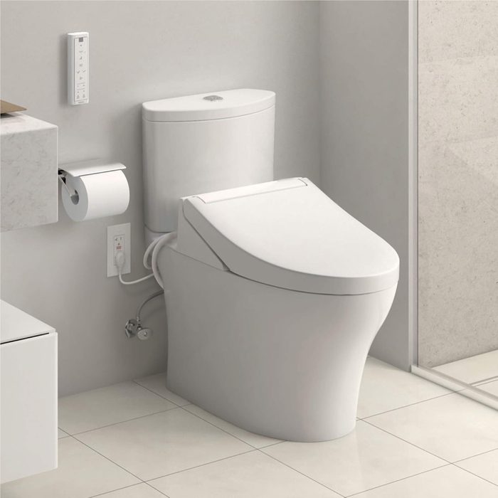 Modern toilet with bidet functionality sits in a minimalist bathroom, surrounded by white tiled floor and light-colored walls.