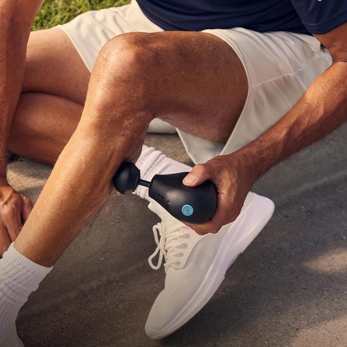 Massage device presses on a man's calf muscle on a sunny outdoor path.