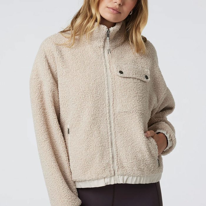 A person wears a beige sherpa jacket with a zipper, hand in pocket, against a plain background.