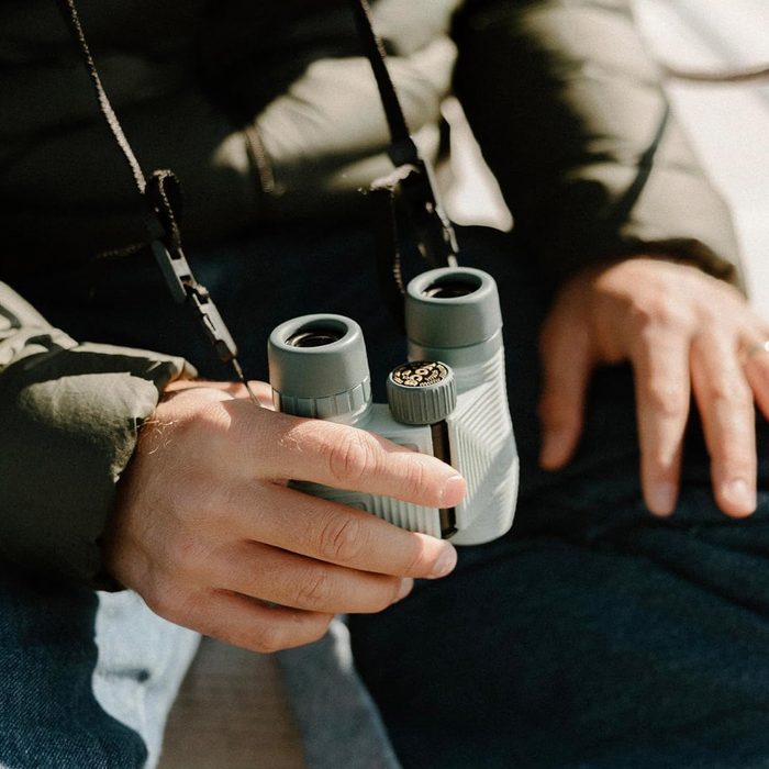 Binoculars held by a hand, resting on a person's lap outdoors, wearing a green jacket and blue jeans.