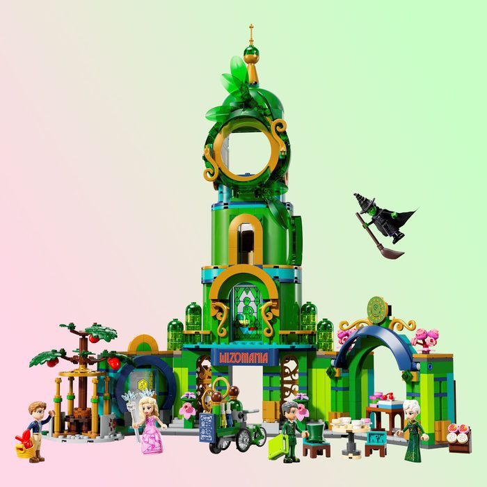 A colorful LEGO set depicts a fantasy scene with figures interacting around a whimsical green tower and garden. A witch flies above on a broom.