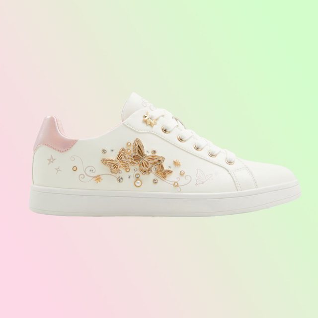 White sneaker features gold butterfly embellishments, displayed against a pastel pink and green gradient backdrop.