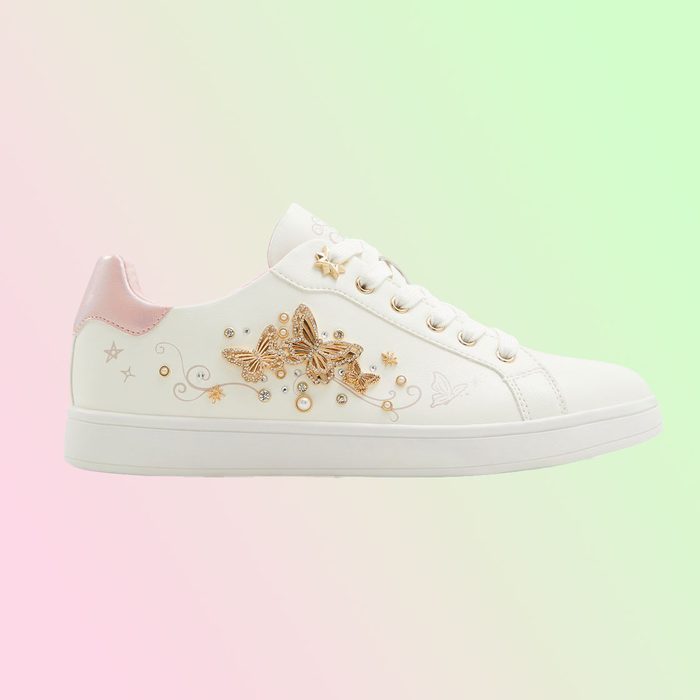 White sneaker features gold butterfly embellishments, displayed against a pastel pink and green gradient backdrop.