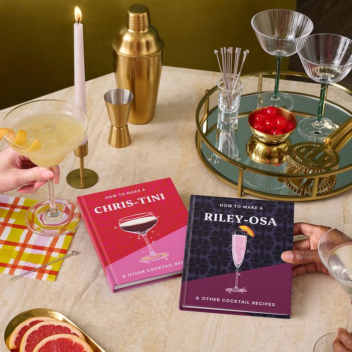 Cocktail books lie on a table. People hold drinks, surrounded by glassware, cherries, and a shaker. A candle provides ambient light.