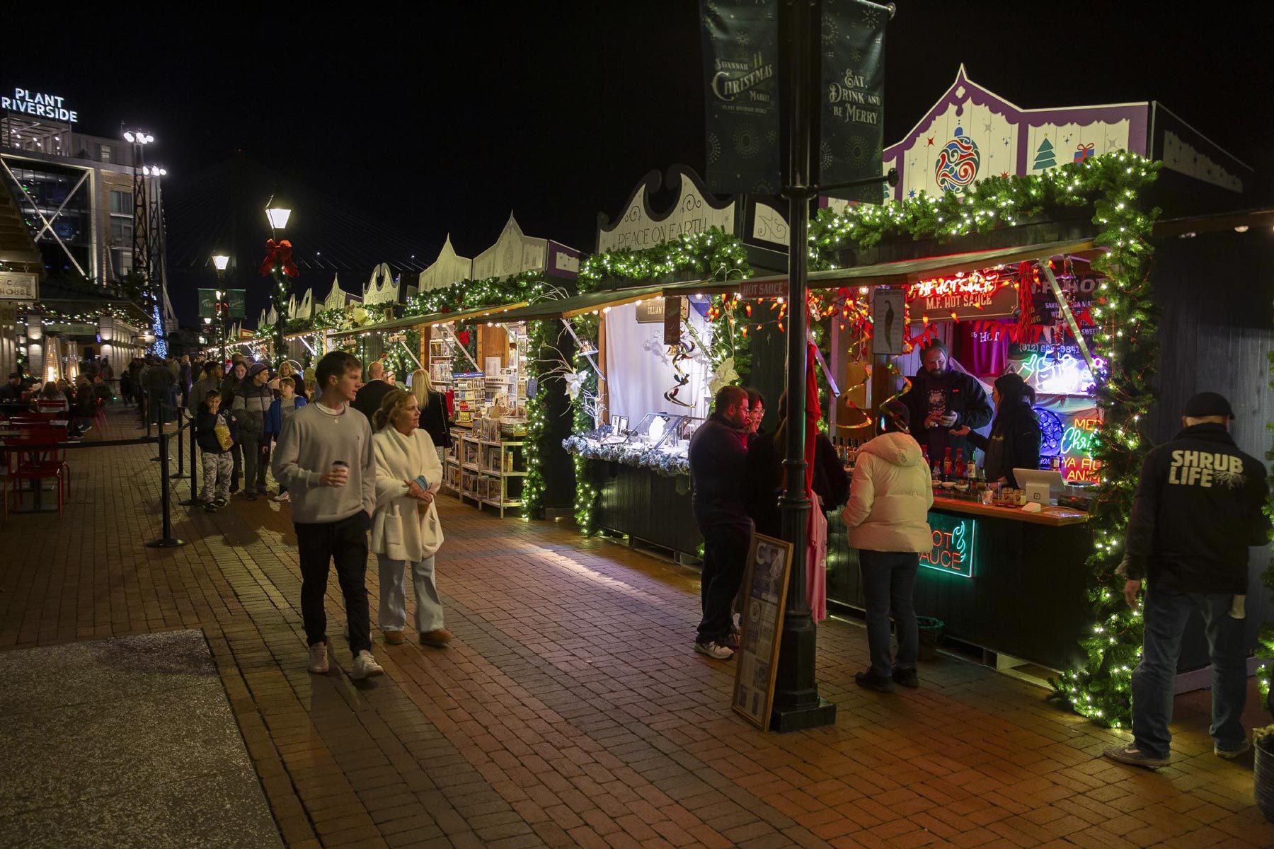 Stalls decorated with lights line a festive night market. People stroll along the brick walkway, browsing and socializing. Signs include "HOT SAUCE" and "Plant Riverside."