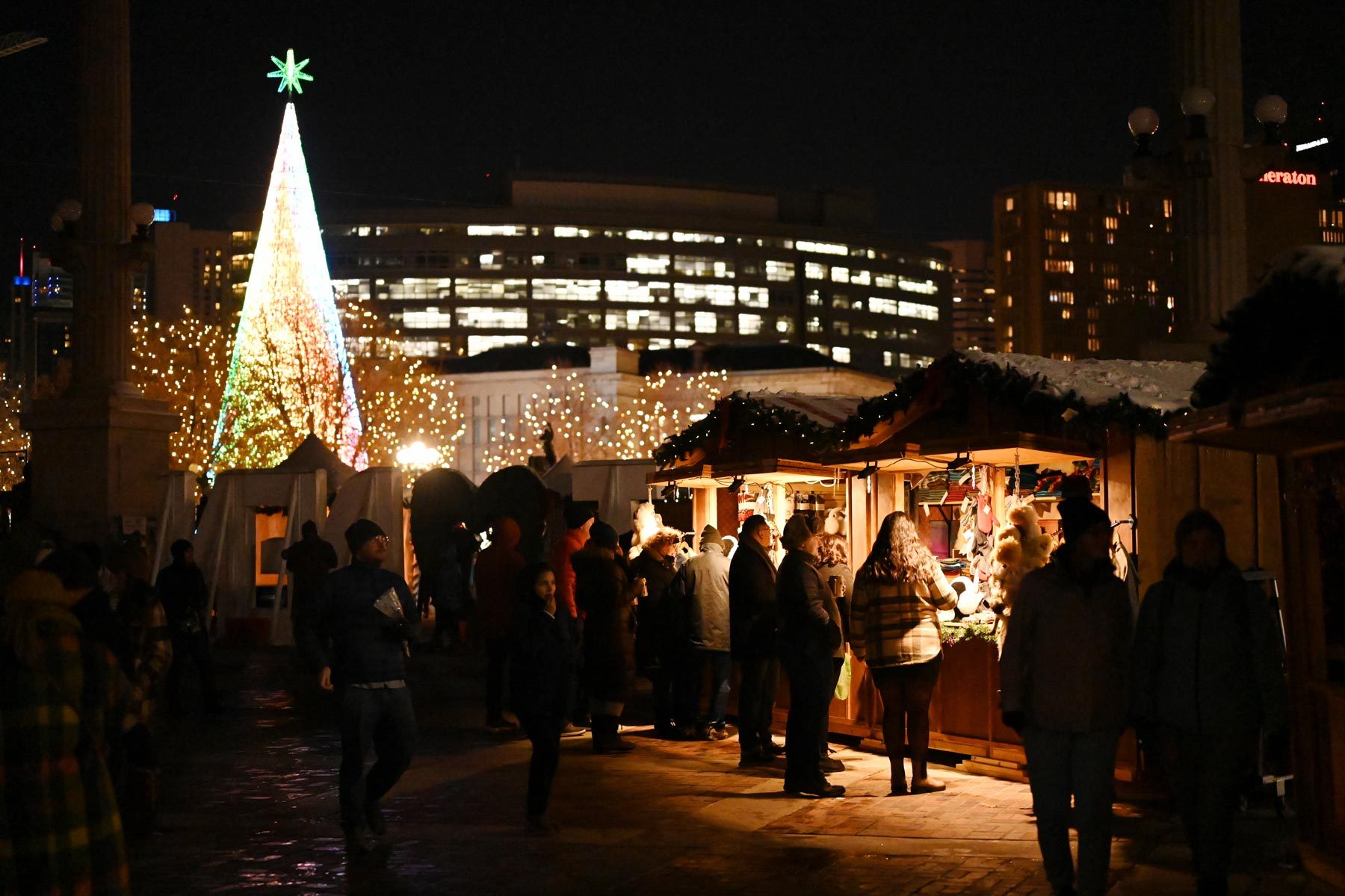 Market stalls illuminated, people shopping leisurely at night; backdrop includes a lit Christmas tree and city buildings adorned with lights.