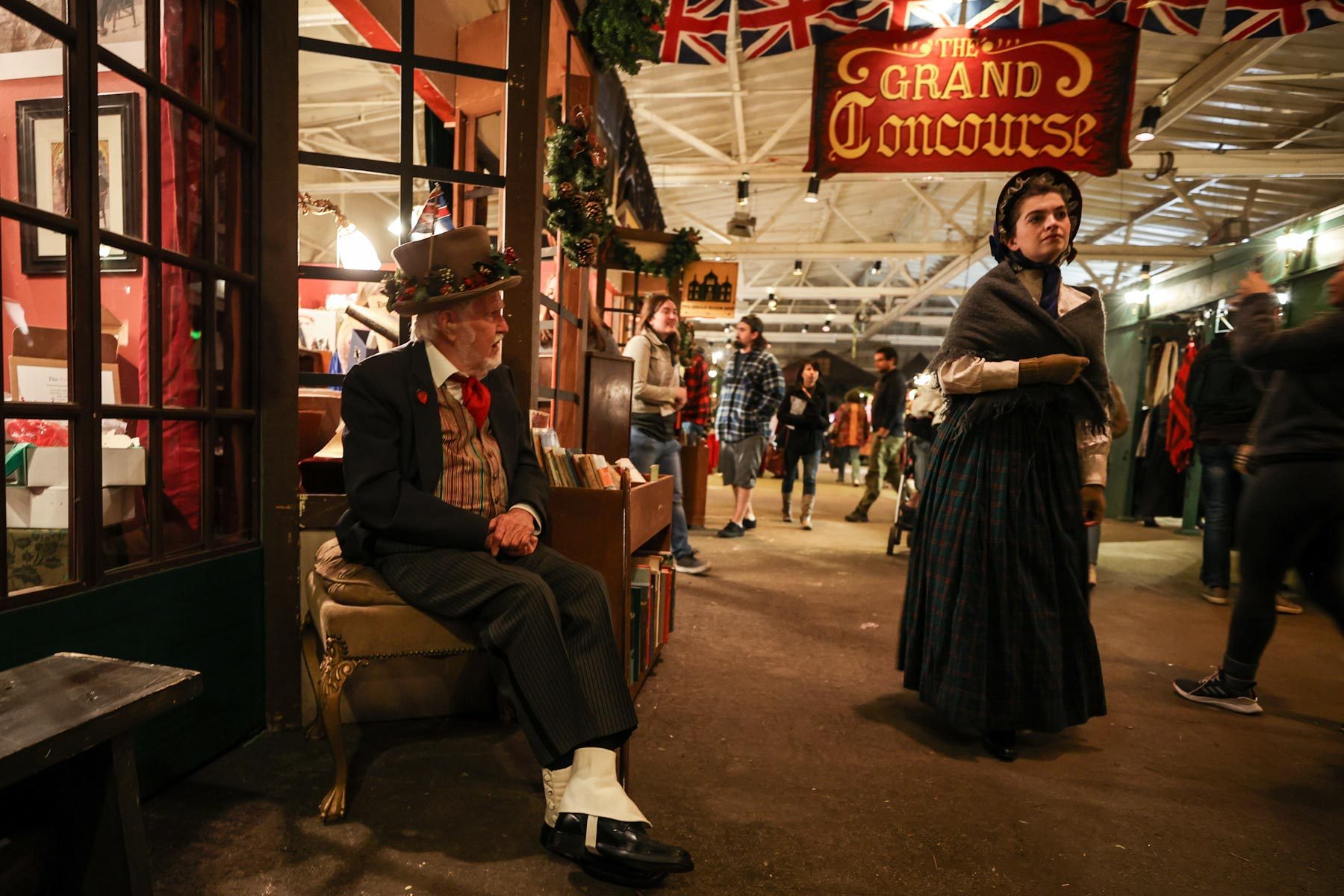 Character sits on a chair, wearing Victorian attire, in a busy indoor marketplace. Overhead sign reads: "The Grand Concourse." People casually walk around.
