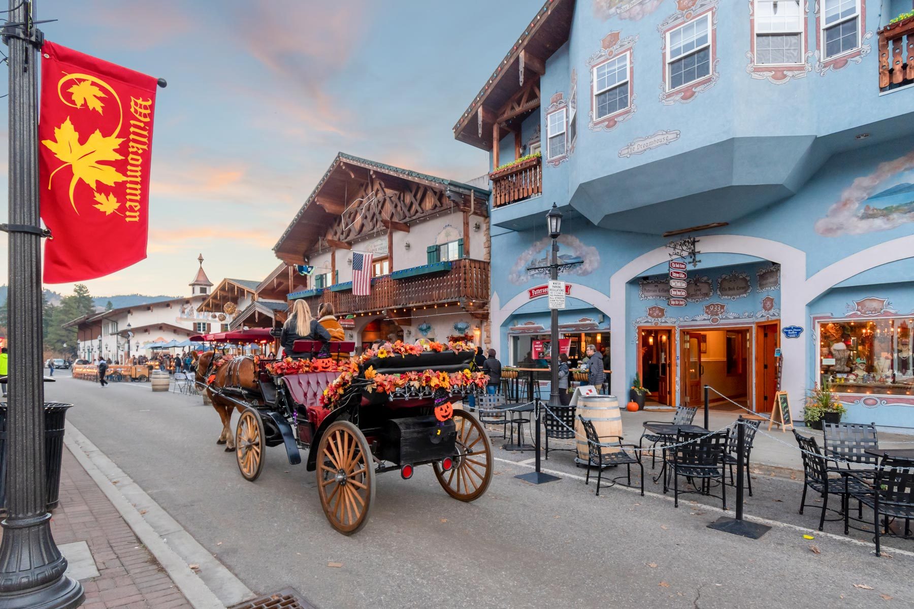 Horse-drawn carriage moves along a festive, Bavarian-style street with ornate buildings and sidewalk dining; red flag reads "Willkommen."