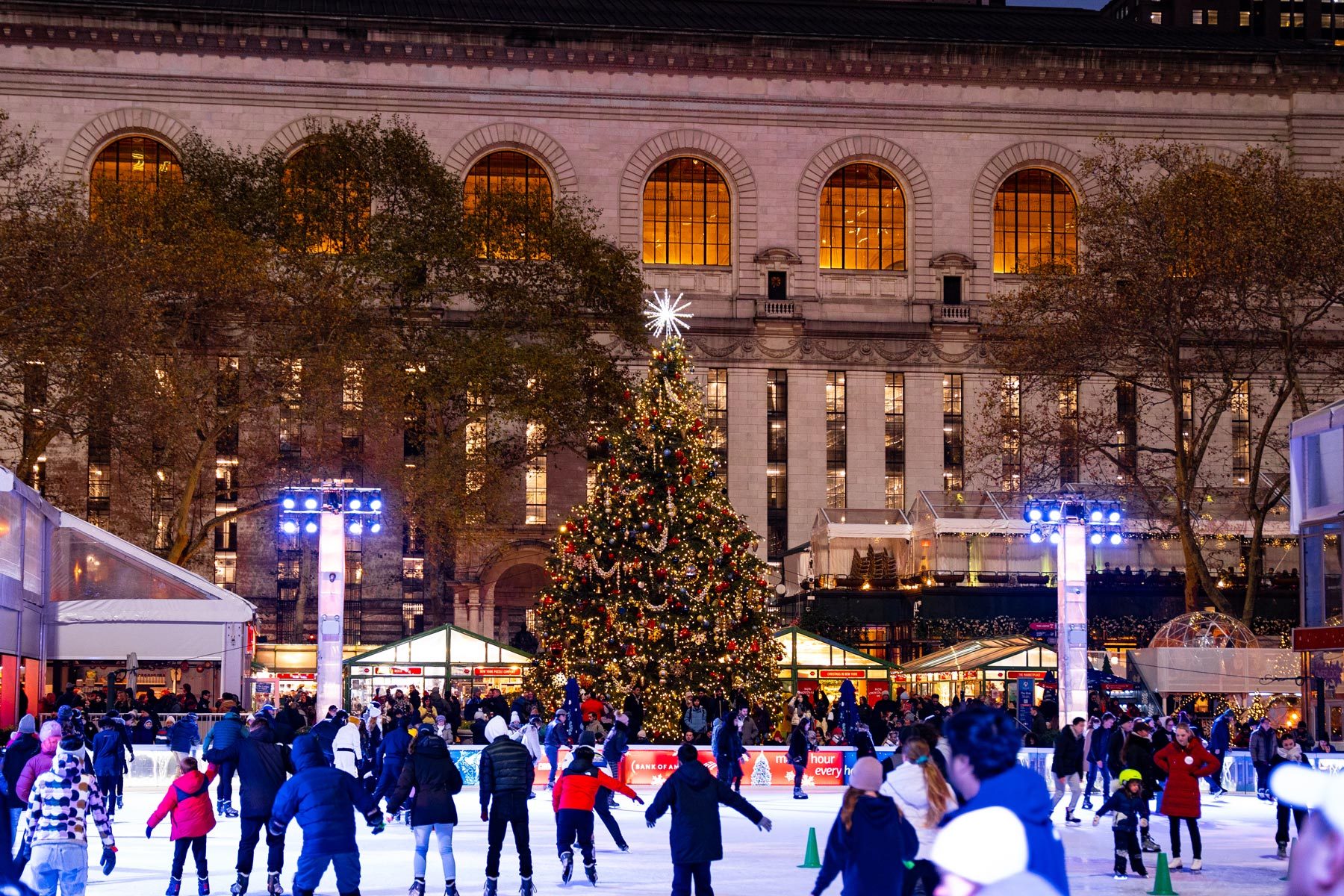Skaters enjoy an ice rink surrounded by festive lights and a decorated Christmas tree, set against the backdrop of a grand building at night.