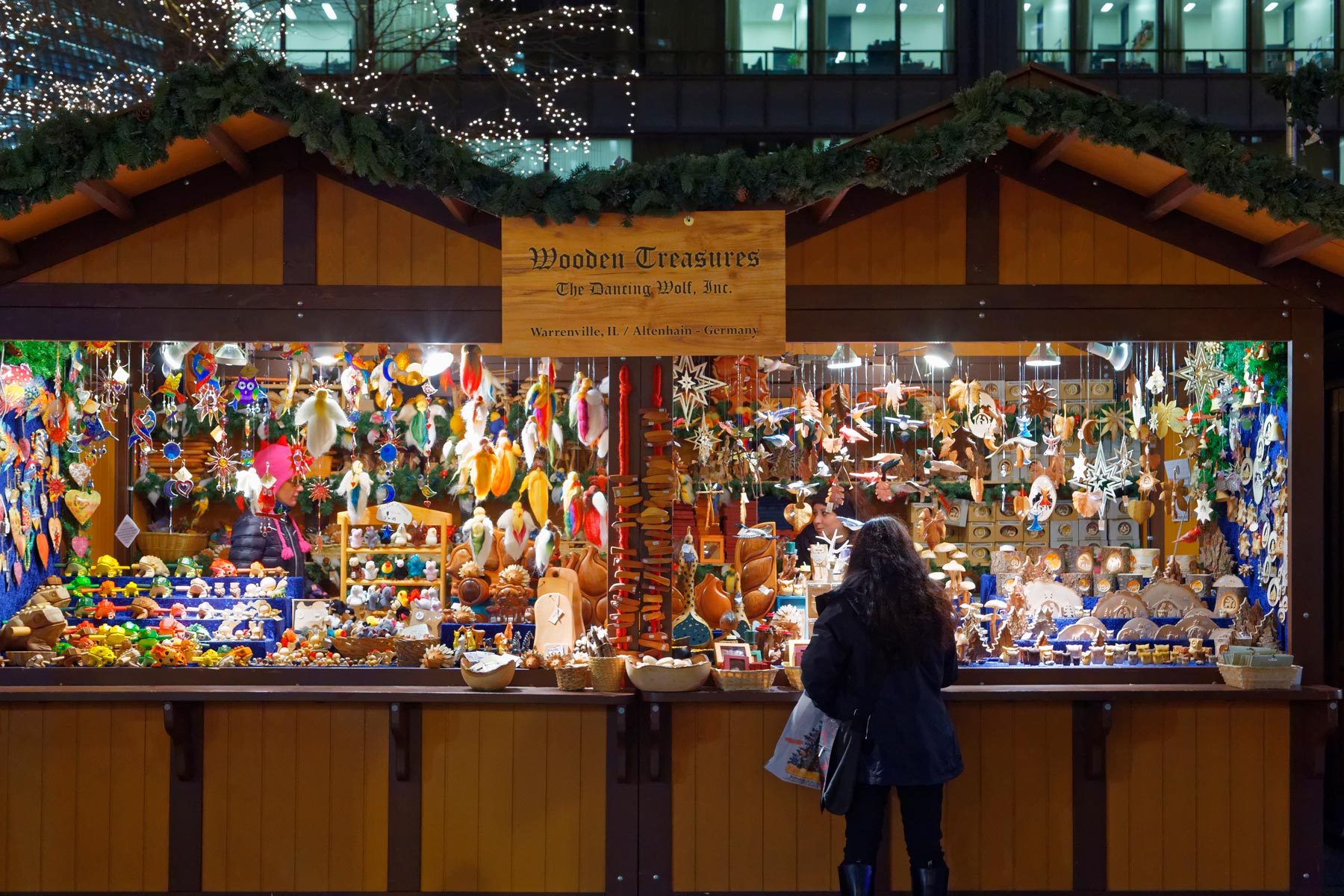 Wooden crafts hang in a festive market stall, while a person examines them. Holiday lights decorate the surrounding night scene.